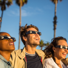 four people wearing eclipse glasses smile while looking into the sky