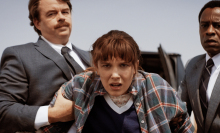 Two men in suits hold the arms of a young woman in a flannel.