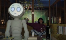 A large white robot with a glowing face and a human woman sit on a couch watching TV.