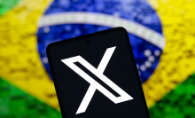 The X (Twitter) logo is displayed on a smartphone screen with a flag of Brazil in the background. Social network X has announced that it will close its office in Brazil.