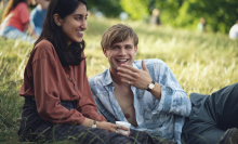 Dexter (Leo Woodall) and Emma (Ambika Mod) are both lying on grass in a park, both laughing. 