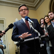 Johnson wears a dark blue suit, white shirt, maroon tie, and spectacles. He stands at a podium/ microphone.