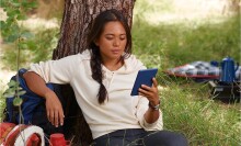 a person sits outside, under a tree while reading on a kindle device