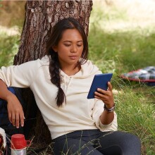 a person sits outside, under a tree while reading on a kindle device