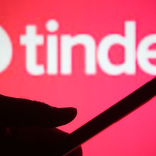 A pink background with Tinder's logo with a silhouette of a hand holding a phone in front of it.