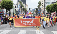 A group of people march down a street during a Pride parade. They are holding a large orange Trevor Project banner. 