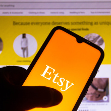 The Etsy logo on a smart phone in front of a desktop screen displaying an Etsy feed.