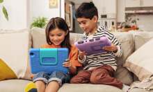 Two kids sitting on the couch and playing on their Amazon Fire 7 Kids tablets with smiles on their faces.