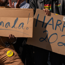 Two people each hold up a cardboard sign. One reads, "Kamala!" The other reads "Harris 2024".