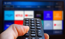 a right hand holds a remote control pointed at a TV with streaming app icons shown on the screen