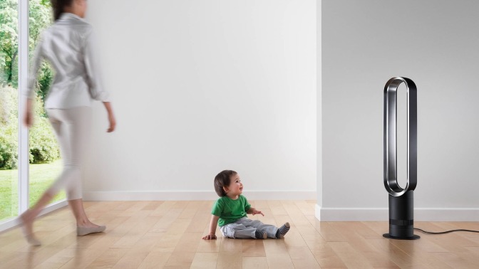 a woman walks across the room to her child who's sitting on the floor in front of a dyson fan