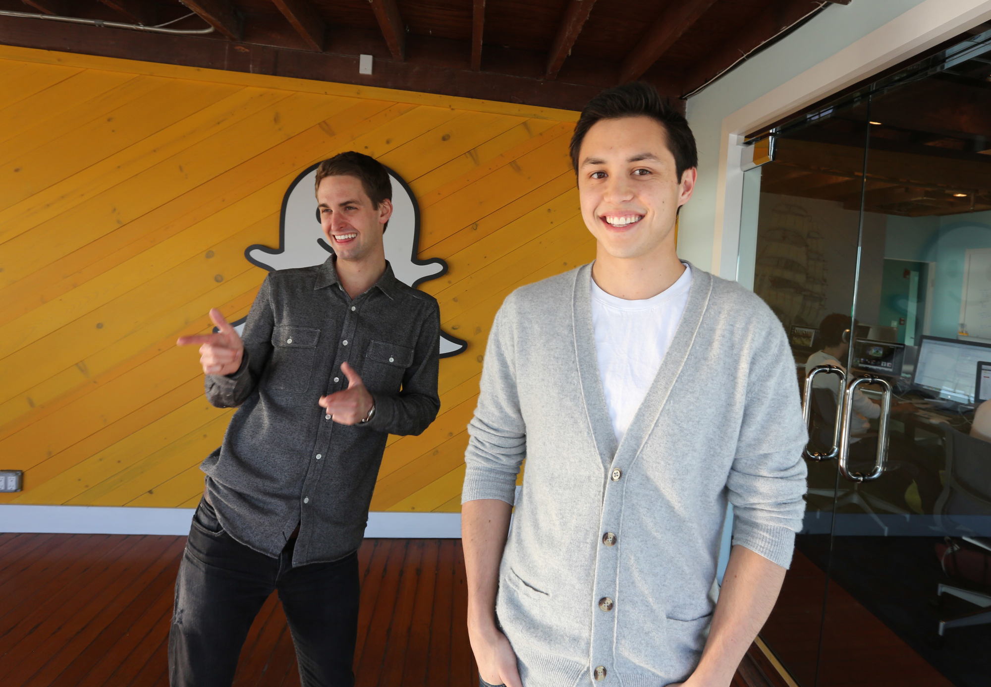 Snapchat co-founders Evan Spiegel and Bobby Murphy smiling and standing in front of a wall decorated with the company's Ghostface Chillah mascot. 