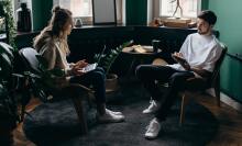 two people are sitting in a corner of a darkly lit room by bay windows, each holding a laptop and iPad air respectively