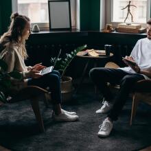 two people are sitting in a corner of a darkly lit room by bay windows, each holding a laptop and iPad air respectively