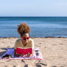 a person lays on a beach towel on the beach reading a red book with the ocean behind her