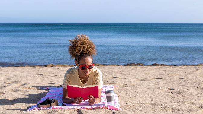 a person lays on a beach towel on the beach reading a red book with the ocean behind her