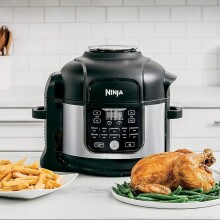 a ninja foodi air fryer and pressure cooker combo sits on a kitchen counter with several dishes of food in front of it including a roast chicken, french fries, and brussel sprouts