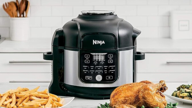 a ninja foodi air fryer and pressure cooker combo sits on a kitchen counter with several dishes of food in front of it including a roast chicken, french fries, and brussel sprouts