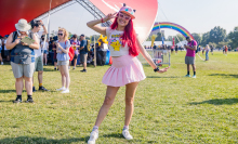 A women in a light pink pleated skirt, white sneakers, and cap. She has red hair and is making a peace sign with her left 