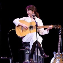 Clairo smiling as she plays the guitar on stage.