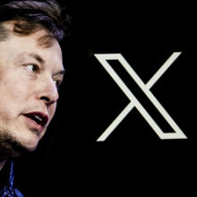 Elon Musk's photo is displayed on a phone screen in front of a computer screen displaying. the Twitter/X logo.