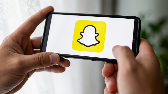 Hands holding a smart phone displaying the yellow and white Snapchat ghost logo. 