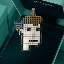 A piece of jewellery in the shape of a pixellated face hangs in front of a green Tiffany box.