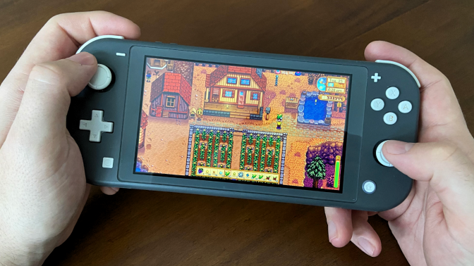 a close-up of a man's hands playing "stardew valley" on a gray nintendo switch lite