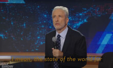 A man on stage in a suit holds a microphone while peering into the audience.