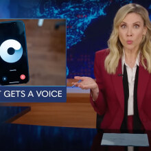 A woman sits behind a talk show desk. In the top-left we can see a smartphone.