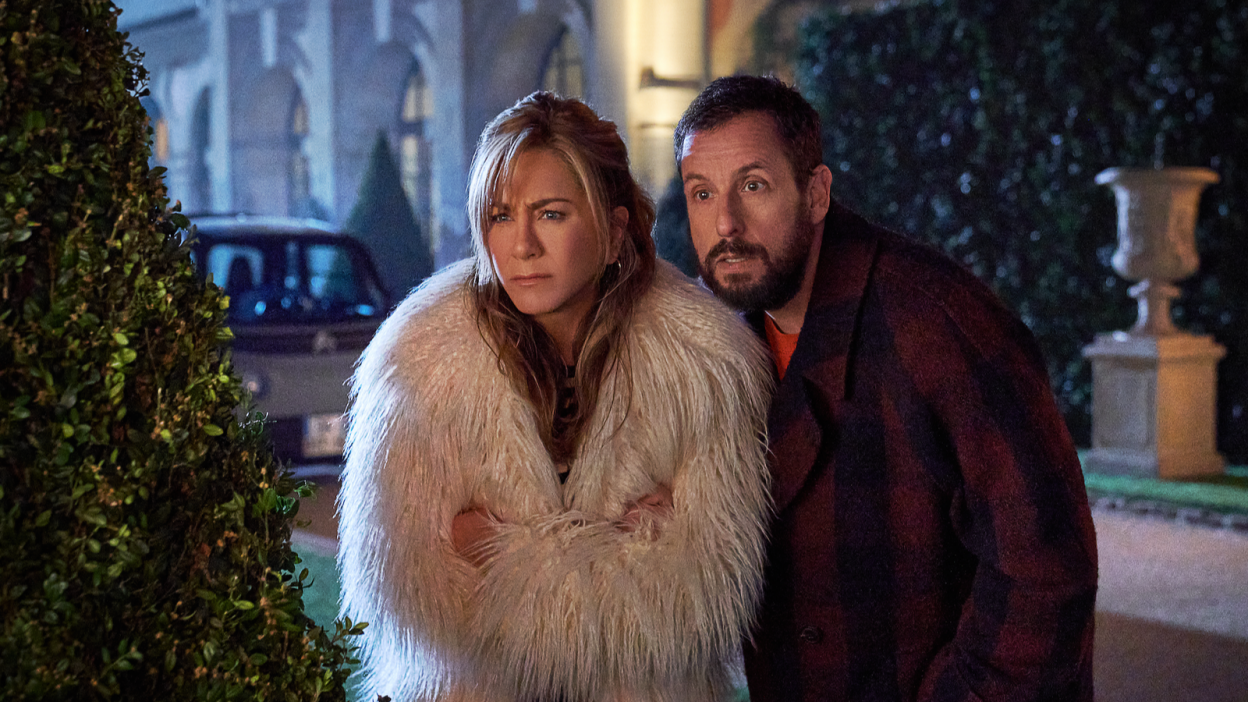 Woman in a fur coat and man in a red trench coat huddle together. 