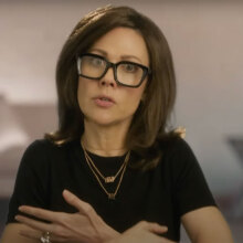 A woman in a brown wig and glasses speaks into the camera.