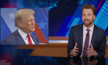 A man sits behind a talk show desk, laughing. In the top-left is an image of another man in a suit.