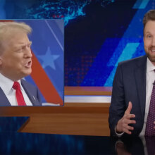 A man sits behind a talk show desk, laughing. In the top-left is an image of another man in a suit.