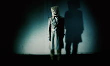 A creepy still from "Emio" showing a person with a bag over their head emblazoned with a smile.