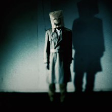 A creepy still from "Emio" showing a person with a bag over their head emblazoned with a smile.