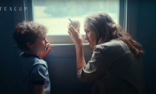 A woman crouches next to a little boy by a window, holding a teacup up to the glass.