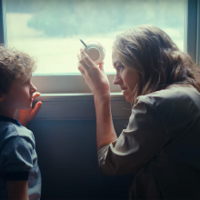 A woman crouches next to a little boy by a window, holding a teacup up to the glass.