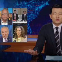 A man in a suit sits behind a talk show desk, with images of six other people in the top left.