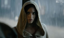 A woman in a grey cloak with the hood up stands indoors, while rain pounds down outside.