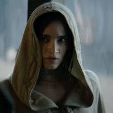 A woman in a grey cloak with the hood up stands indoors, while rain pounds down outside.