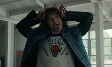 A young man in a shirt with a devil on it makes devil horns above his head.