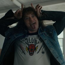 A young man in a shirt with a devil on it makes devil horns above his head.