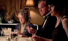 Three characters from 'Downton Abbey' sit at a dinner table. Maggie Smith's character, Dowager Countess, looks at the camera.