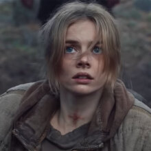 A woman sits on a chair in the woods, looking afraid.