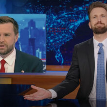 A man in a suit sits behind a talk show desk with his arms spread wide. In the top-left is an image of another man in a suit.