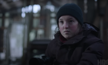 A teen girl wearing a woollen hat stands in a forest covered in snow, looking serious.