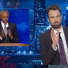 A man sits behind a talk show desk. In the top-left is an image of another man speaking at a podium.