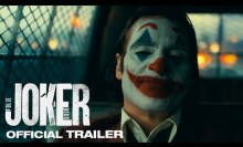 Joker sits in the back of a police car, with his full face of clown makeup on.