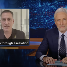 A man in a suit sits behind a talk show desk. In the top-left is an image of a man on the news and the phrase "de-escalation through escalation."